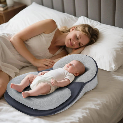 Bébé allongé dans un cocon pour bebe gris et bleu marine sur un lit blanc, accompagné de sa maman dans une chambre paisible et chaleureuse.