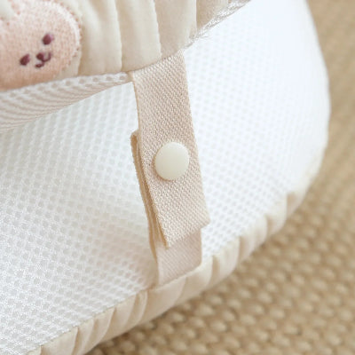 Reducteur de lit bebe gros plan sur une attache pression beige fixĂ©e Ă une maille respirante blanche, bordĂ©e dâun tissu doux. DĂ©tail de finition soignĂ©e, illustrant la sĂ©curitĂ© et le confort du cocon.