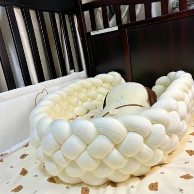Tour de lit tressé : tresse blanche moelleuse entourant un coussin en forme de chien, posée sur drap crÚme à motifs oursons dans un berceau aux barreaux foncés. Confort et sécurité pour bébé.
