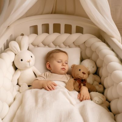 bébé de 12 mois endormi dans berceau blanc, sur oreiller et sous couverture, entouré de peluches et d’un tour de lit tressé blanc, sous un ciel de lit dans une chambre chaleureuse.