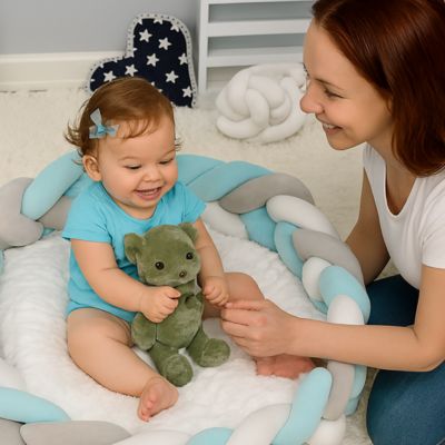 Tresse de lit bébé : bébé joue avec sa doudou dans un cocon tressé pastel bleu, gris et blanc, accompagné de sa maman. Moment doux et complice.