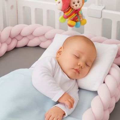 Tresse de lit bébé : bébé de 6 mois endormi au centre d’un berceau blanc avec tresse rose tendre, oreiller moelleux, couverture douce et peluches suspendues. Ambiance cocooning.