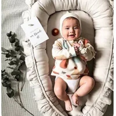 un bebe souriant qui est allonger dans son nid pour bebe couleur gris sur un matelas blanc a cote de branche. 