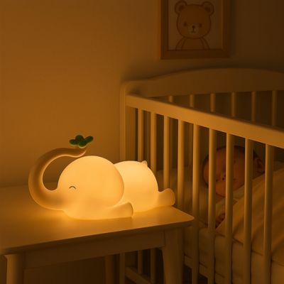 veilleuse éléphant allumé sur une table de nuit a cote d'un berceau avec un bébé qui dors paisiblement dedans et un cadre de photo dessus 